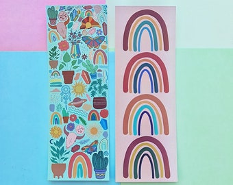 Puede incluir: Dos pegatinas rectangulares. Una es de color verde azulado claro con ilustraciones coloridas de plantas, cristales y una mariposa. La otra es de color rosa claro con cuatro diseños de arcoíris en tonos marrón, azul y amarillo. El fondo es una mezcla de rosa, azul y verde.