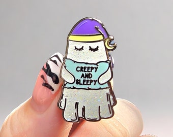May include: A whimsical enamel pin featuring a cartoon ghost wearing a purple nightcap with a crescent moon. The ghost is holding a light blue shirt with the words "Creepy and Sleepy". The ghost has closed eyes and a glittery skirt.