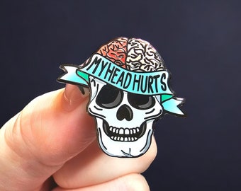 May include: A skull-shaped enamel pin with a brain design and the words "MY HEAD HURTS" on a teal banner. The skull is white with black eye sockets and a detailed jawline. The brain is colored in shades of red and tan.
