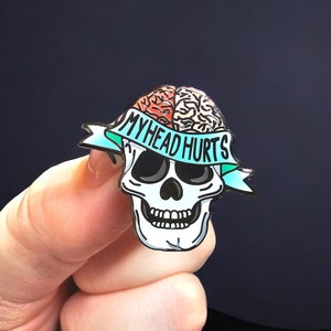 Könnte beinhalten: Ein schädelformiger Emaille-Pin mit einem Gehirndesign und dem Schriftzug "MY HEAD HURTS" auf einem türkisfarbenen Banner. Der Schädel ist weiß mit schwarzen Augenhöhlen und einem detaillierten Kiefer. Das Gehirn ist in Rot- und Beigetönen gehalten.