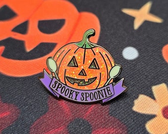 May include: A Halloween-themed enamel pin featuring an orange pumpkin with a smiling face and glitter accents. The pin includes two spoons and a purple banner that reads "Spooky Spoonie". The pin is set against a black background with orange pumpkins and floral patterns.