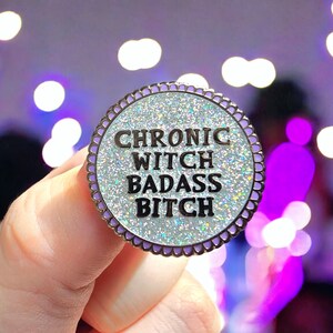 Puede incluir: Un pin redondo de esmalte con el texto "CHRONIC WITCH BADASS BITCH" en letras negras. El pin tiene un centro plateado brillante, un borde lavanda y un borde festoneado. El fondo está borroso con luces moradas y blancas.
