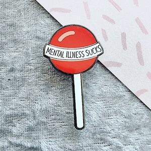 May include: A red lollipop-shaped enamel pin with the text "MENTAL ILLNESS SUCKS" on a white banner. The pin has a white stick and is set against a gray textured background.