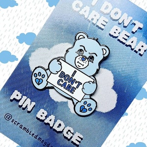 Peut inclure: Épinglette en émail bleu clair en forme d'ours tenant une pancarte avec l'inscription « I DON'T CARE ». L'épingle est posée sur un fond bleu avec les mots « I DON'T CARE BEAR » et « PIN BADGE ». L'épingle a des contours et des détails noirs.