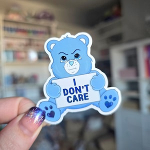 May include: A blue cartoon bear sticker with an angry expression, holding a sign that reads "I DON'T CARE" in bold, blue letters. The bear has a heart-shaped nose and heart-shaped paw prints. The sticker is on a white background.