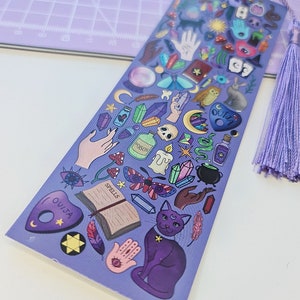 May include: A purple bookmark with a tassel, featuring various mystical illustrations. The design includes a cat, Ouija board, crystals, potion bottles, and a spell book. The bookmark is decorated with a variety of colors and shapes.