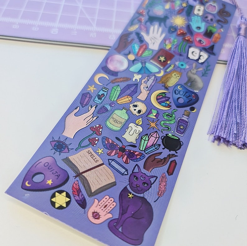 May include: A purple bookmark with a tassel, featuring various mystical illustrations. The design includes a cat, Ouija board, crystals, potion bottles, and a spell book. The bookmark is decorated with a variety of colors and shapes.