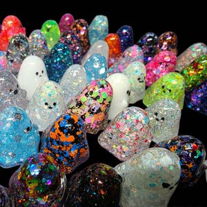 Op de afbeelding: Een verzameling kleine, afgeronde spookvormige beeldjes gemaakt van hars. Elk spookje heeft een unieke afwerking, waaronder diverse veelkleurige glitters, iriserende vlokken, effen dekkend wit en doorschijnende texturen, allemaal met eenvoudige zwarte oogjes.