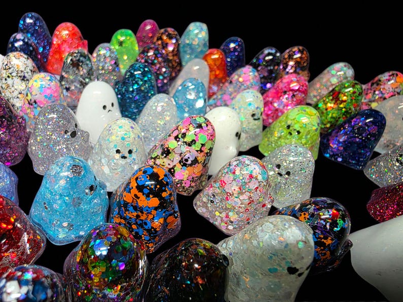 Op de afbeelding: Een verzameling kleine, afgeronde spookvormige beeldjes gemaakt van hars. Elk spookje heeft een unieke afwerking, waaronder diverse veelkleurige glitters, iriserende vlokken, effen dekkend wit en doorschijnende texturen, allemaal met eenvoudige zwarte oogjes.