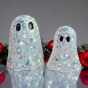 May include: Two glittery white ghost figurines with black eyes. The ghosts are decorated with iridescent glitter and are surrounded by red Christmas ornaments and green pine needles. The ghosts are a festive holiday decoration.
