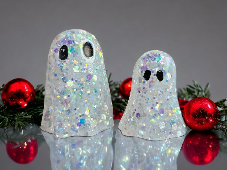 May include: Two glittery white ghost figurines with black eyes. The ghosts are decorated with iridescent glitter and are surrounded by red Christmas ornaments and green pine needles. The ghosts are a festive holiday decoration.