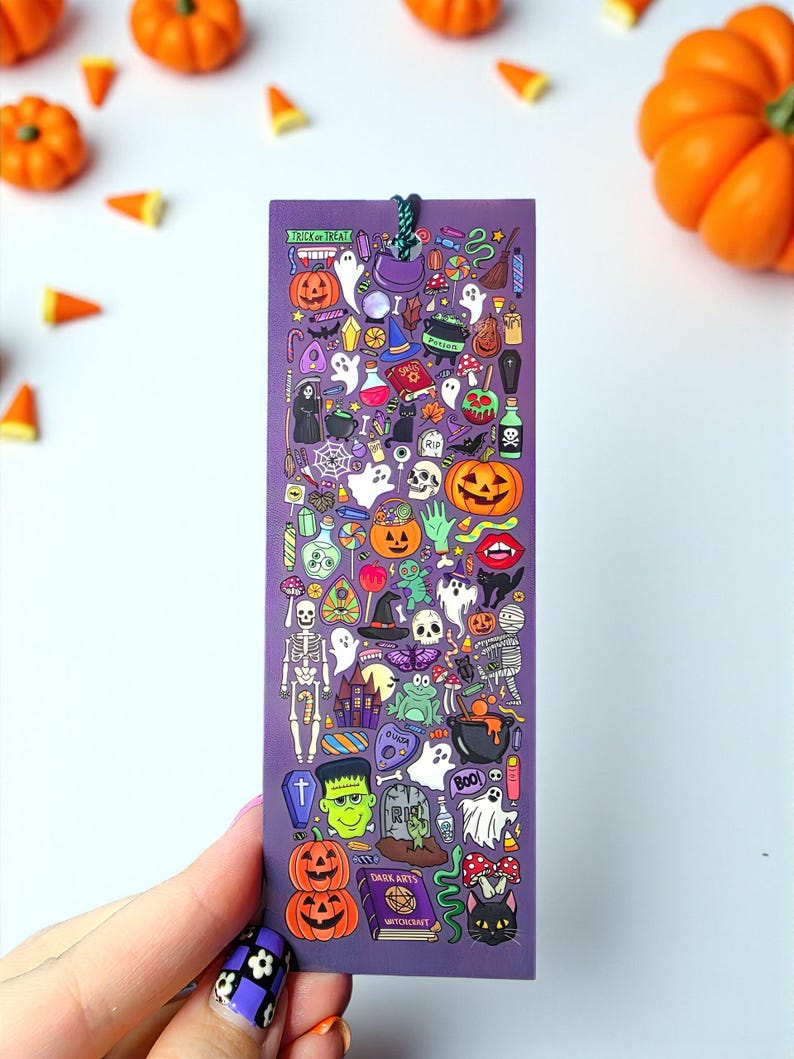 Puede incluir: Un marcap&aacute;ginas morado con ilustraciones de Halloween, incluyendo calabazas, fantasmas y sombreros de bruja. El marcap&aacute;ginas tiene una cuerda verde y las palabras "Trick or Treat". Calabazas naranjas y caramelos est&aacute;n esparcidos.