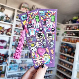 Pode incluir: Dois marcadores de livros roxos com uma borla rosa, com ilustra&ccedil;&otilde;es tem&aacute;ticas dos anos 90. Os designs incluem um Cubo de Rubik, uma fita cassete, um unic&oacute;rnio, Furby, computador, Game Boy e um cora&ccedil;&atilde;o Polly Pocket.