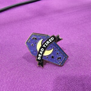 May include: A purple and gold enamel pin in the shape of a coffin, with a crescent moon and stars design. The pin has a banner that reads "DEAD TIRED" in white text. The pin is set against a purple background.