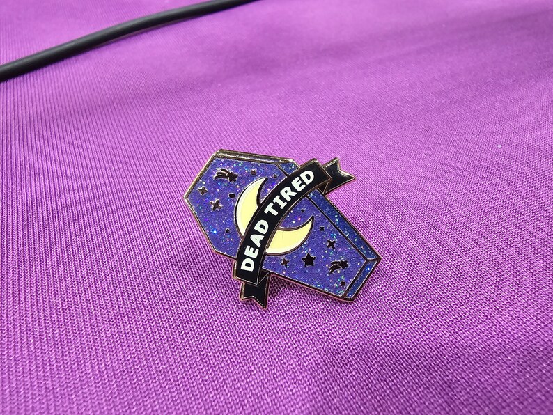 May include: A purple and gold enamel pin in the shape of a coffin, with a crescent moon and stars design. The pin has a banner that reads "DEAD TIRED" in white text. The pin is set against a purple background.