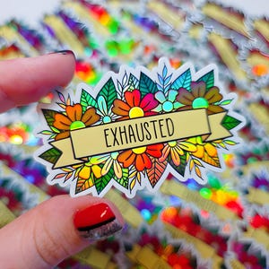 May include: A colorful sticker featuring a floral design with the word "EXHAUSTED" on a banner. The sticker has a holographic background and is held by a hand. The flowers are in shades of orange, red, yellow, and green.