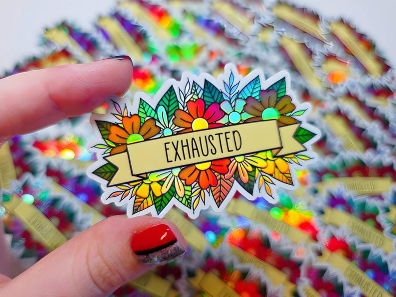 May include: A colorful sticker featuring a floral design with the word "EXHAUSTED" on a banner. The sticker has a holographic background and is held by a hand. The flowers are in shades of orange, red, yellow, and green.