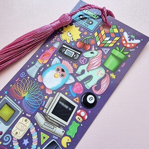 Pode incluir: Marcador de livros roxo com borla rosa, com ilustra&ccedil;&otilde;es tem&aacute;ticas dos anos 90. Inclui um Furby, Lisa Simpson, um cubo de Rubik e uma fita VHS. O marcador tem um furo na parte superior para a borla.
