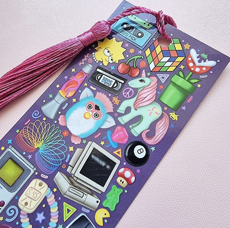 Pode incluir: Marcador de livros roxo com borla rosa, com ilustra&ccedil;&otilde;es tem&aacute;ticas dos anos 90. Inclui um Furby, Lisa Simpson, um cubo de Rubik e uma fita VHS. O marcador tem um furo na parte superior para a borla.