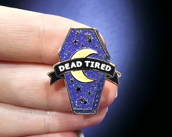 May include: A coffin-shaped enamel pin with a glittery blue background, a yellow crescent moon, and stars. The pin has a black banner that reads "DEAD TIRED" in white lettering. The pin has gold-colored trim.