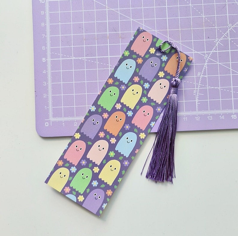 May include: A colorful bookmark featuring a pattern of smiling ghost figures in various pastel colors, set against a dark purple background with small flower accents. A matching purple tassel hangs from the top. A cute and whimsical design.