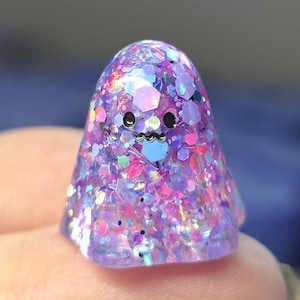 Op de afbeelding: Een klein, koepelvormig spookfiguurtje gemaakt van doorschijnende paarse hars. Het oppervlak is ingelegd met een dichte mix van iriserende, holografische en zeshoekige glitters in roze, blauwe en paarse tinten. Het heeft twee zwarte ogen.