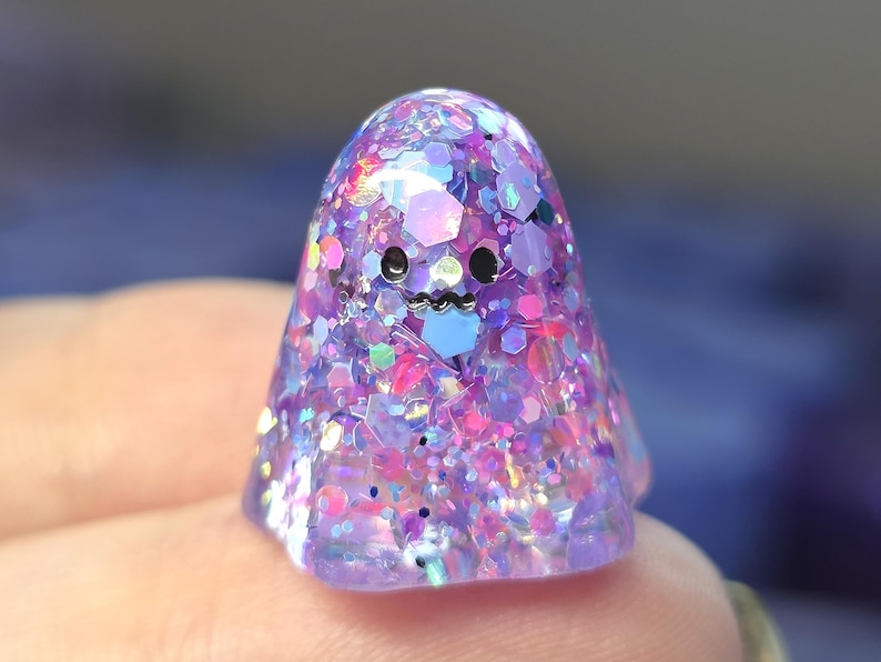 Op de afbeelding: Een klein, koepelvormig spookfiguurtje gemaakt van doorschijnende paarse hars. Het oppervlak is ingelegd met een dichte mix van iriserende, holografische en zeshoekige glitters in roze, blauwe en paarse tinten. Het heeft twee zwarte ogen.