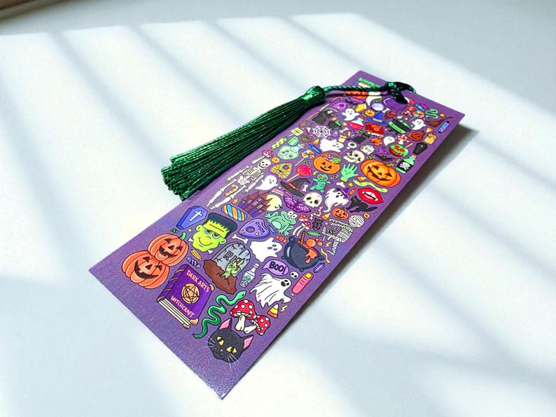 Puede incluir: Un marcap&aacute;ginas morado con tem&aacute;tica de Halloween y borla verde. El marcap&aacute;ginas est&aacute; cubierto de ilustraciones coloridas de calabazas, fantasmas, Frankenstein y otras im&aacute;genes espeluznantes. Las palabras "DARK ARTS" y "WITCHCRAFT" son visibles en un libro.