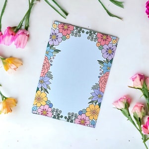 May include: A pastel blue notepad featuring a floral border in pink, yellow, and purple. The notepad is surrounded by fresh flowers in shades of pink, yellow, and purple, creating a cheerful and artistic composition.