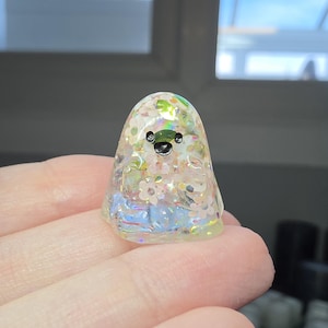 Op de afbeelding: Een klein, doorschijnend, geestvormig beeldje gemaakt van hars. Het figuurtje heeft een iriserende afwerking met ingebedde witte bloemvormige glitters en kleine, glinsterende lovertjes. Het heeft twee kleine zwarte stippen als ogen en een mond.