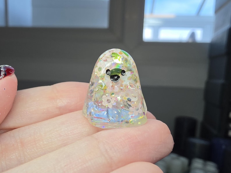 Op de afbeelding: Een klein, doorschijnend, geestvormig beeldje gemaakt van hars. Het figuurtje heeft een iriserende afwerking met ingebedde witte bloemvormige glitters en kleine, glinsterende lovertjes. Het heeft twee kleine zwarte stippen als ogen en een mond.