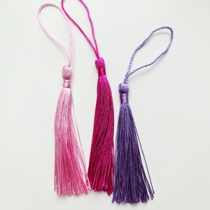 May include: Three colorful tassels in shades of pink and purple. Each tassel has a loop at the top and a tightly wound section above the flowing strands. The tassels are arranged against a white background.