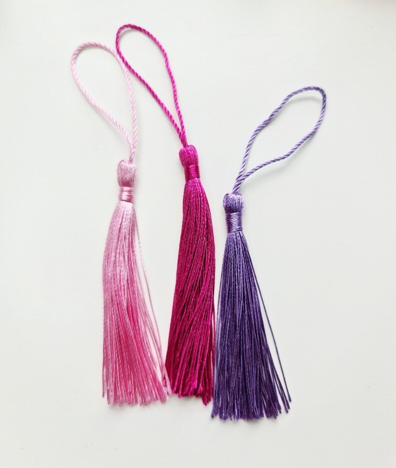 May include: Three colorful tassels in shades of pink and purple. Each tassel has a loop at the top and a tightly wound section above the flowing strands. The tassels are arranged against a white background.