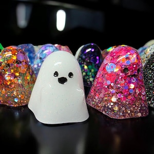 Op de afbeelding: Een verzameling kleine, spookvormige harsfiguurtjes in diverse kleuren en afwerkingen. De spoken hebben eenvoudige zwarte stippen als ogen en monden, sommige gevuld met grove iriserende glitters, andere in effen wit of donkere glitters.
