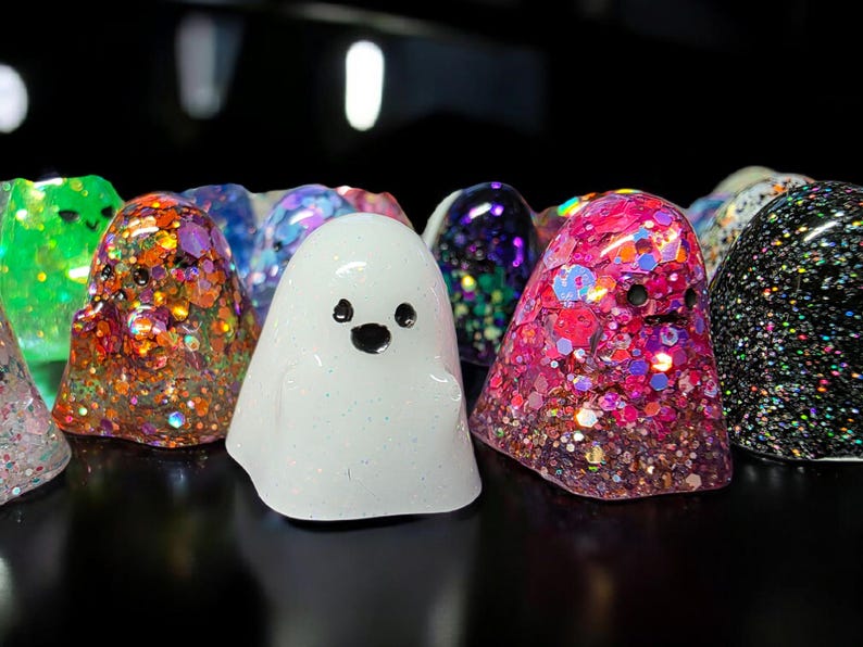 Op de afbeelding: Een verzameling kleine, spookvormige harsfiguurtjes in diverse kleuren en afwerkingen. De spoken hebben eenvoudige zwarte stippen als ogen en monden, sommige gevuld met grove iriserende glitters, andere in effen wit of donkere glitters.