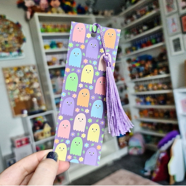May include: A colorful bookmark featuring a pattern of smiling ghost characters in pastel colors on a purple background, with small flowers. The bookmark has a matching purple tassel attached at the top, perfect for book lovers.