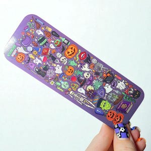 Puede incluir: Un marcap&aacute;ginas morado con tem&aacute;tica de Halloween, cubierto de ilustraciones de calabazas, fantasmas, esqueletos y otras im&aacute;genes espeluznantes. El marcap&aacute;ginas es rectangular con esquinas redondeadas.