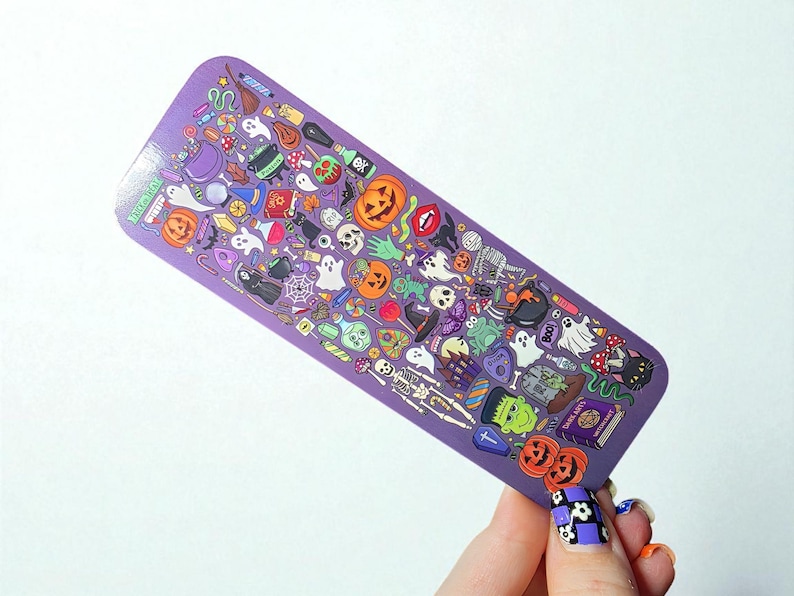 Puede incluir: Un marcap&aacute;ginas morado con tem&aacute;tica de Halloween, cubierto de ilustraciones de calabazas, fantasmas, esqueletos y otras im&aacute;genes espeluznantes. El marcap&aacute;ginas es rectangular con esquinas redondeadas.