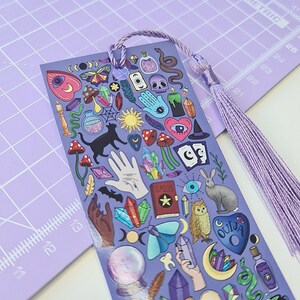 May include: A purple bookmark with a tassel, featuring various mystical illustrations. The design includes a black cat, mushrooms, crystals, a hand, an owl, and a Ouija board. The bookmark is made of a glossy material.