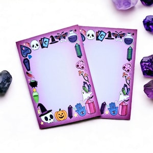 May include: Two rectangular purple notepads with a border of Halloween-themed illustrations, including skulls, potions, and crystals. The notepads are surrounded by purple crystals and a dark gray stone on a white surface.