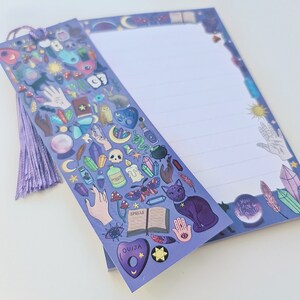 May include: A lavender-colored notepad and bookmark set with a mystical theme. The bookmark features various occult symbols, a purple tassel, and the word "Spells" on a book. The notepad has lined pages and a border with celestial and crystal illustrations.