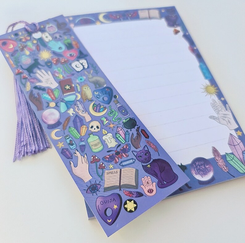 May include: A lavender-colored notepad and bookmark set with a mystical theme. The bookmark features various occult symbols, a purple tassel, and the word "Spells" on a book. The notepad has lined pages and a border with celestial and crystal illustrations.