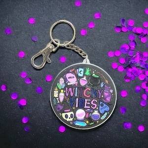 May include: A circular keychain with the text "Witchy Vibes" in pink and purple, surrounded by illustrations of spells, potions, a cat, and a skull. It has a silver clasp and chain. Purple confetti is scattered around the keychain.