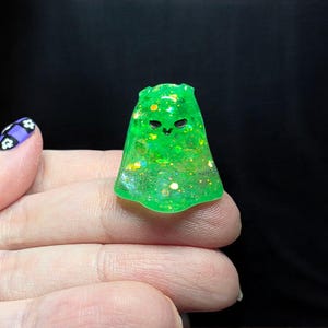 Op de afbeelding: Een klein, doorschijnend neon-groen spookvormig figuurtje gemaakt van hars. Het oppervlak is ingelegd met diverse iriserende glittervlokken die het licht vangen. Het heeft twee eenvoudige zwarte ogen en een kleine mond voor een vriendelijke uitstraling.