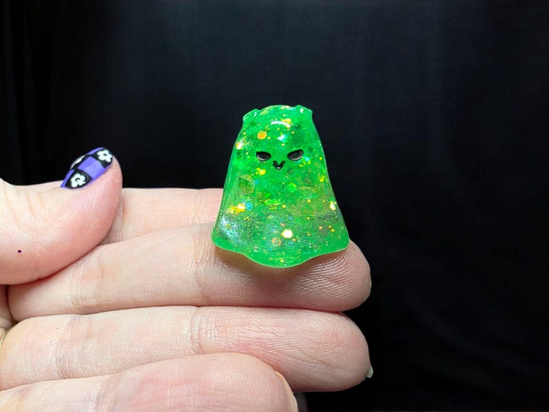 Op de afbeelding: Een klein, doorschijnend neon-groen spookvormig figuurtje gemaakt van hars. Het oppervlak is ingelegd met diverse iriserende glittervlokken die het licht vangen. Het heeft twee eenvoudige zwarte ogen en een kleine mond voor een vriendelijke uitstraling.
