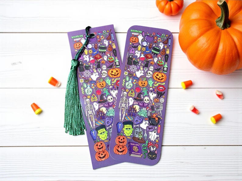 Puede incluir: Dos marcap&aacute;ginas morados con tem&aacute;tica de Halloween con borlas verdes, con ilustraciones de calabazas, fantasmas y otros elementos espeluznantes. Caramelos y calabazas est&aacute;n esparcidos sobre una superficie de madera blanca.