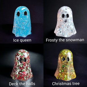 May include: Four resin ghost figurines with black eyes. The ghosts are labeled "Ice queen", "Frosty the snowman", "Deck the halls", and "Christmas tree". Each ghost is filled with glitter and decorative elements in various colors.