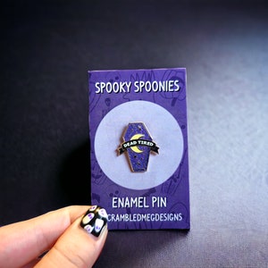 May include: A purple enamel pin in the shape of a coffin with a crescent moon and stars design. The pin has the words "Dead Tired" on a black banner. The pin is on a purple card with the words "Spooky Spoonies" and "Enamel Pin".