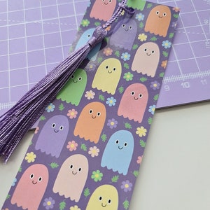 May include: A colorful bookmark featuring a pattern of smiling ghost characters in pastel colors on a purple background. The bookmark has a matching purple tassel attached to the top. The design includes small flowers.