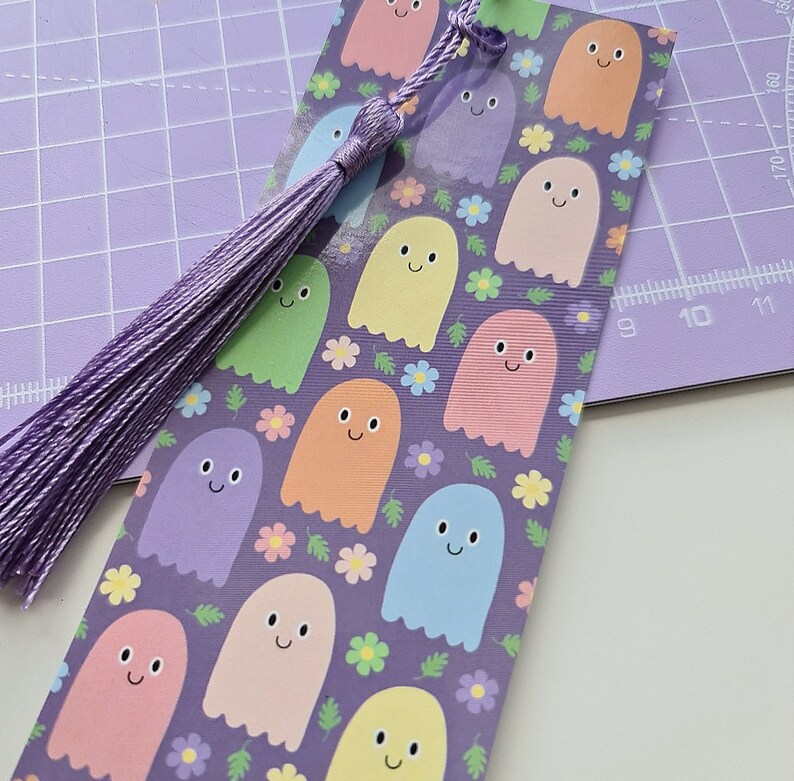 May include: A colorful bookmark featuring a pattern of smiling ghost characters in pastel colors on a purple background. The bookmark has a matching purple tassel attached to the top. The design includes small flowers.